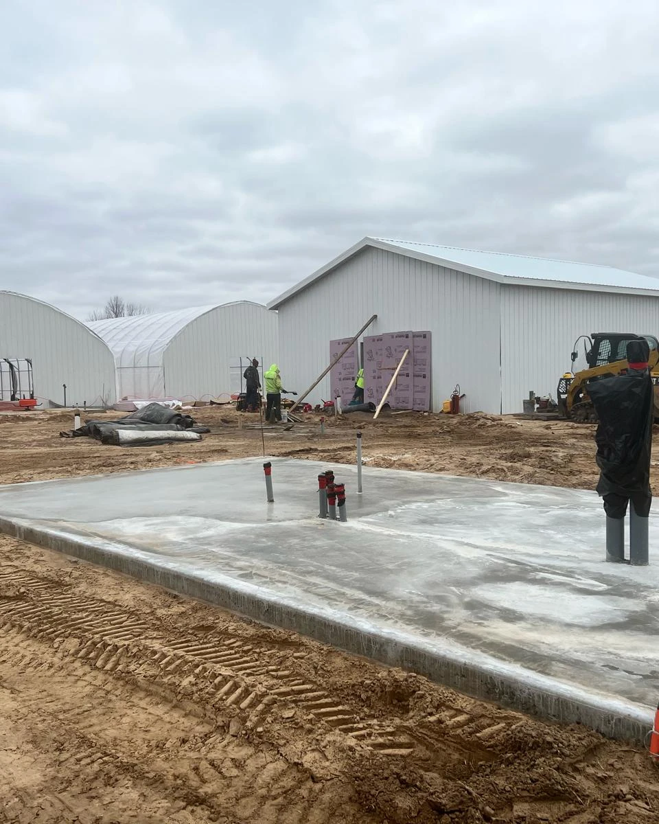 The Michigan site under construction: a freshly poured concrete slab in front of two greenhouse frames and a steel-clad building